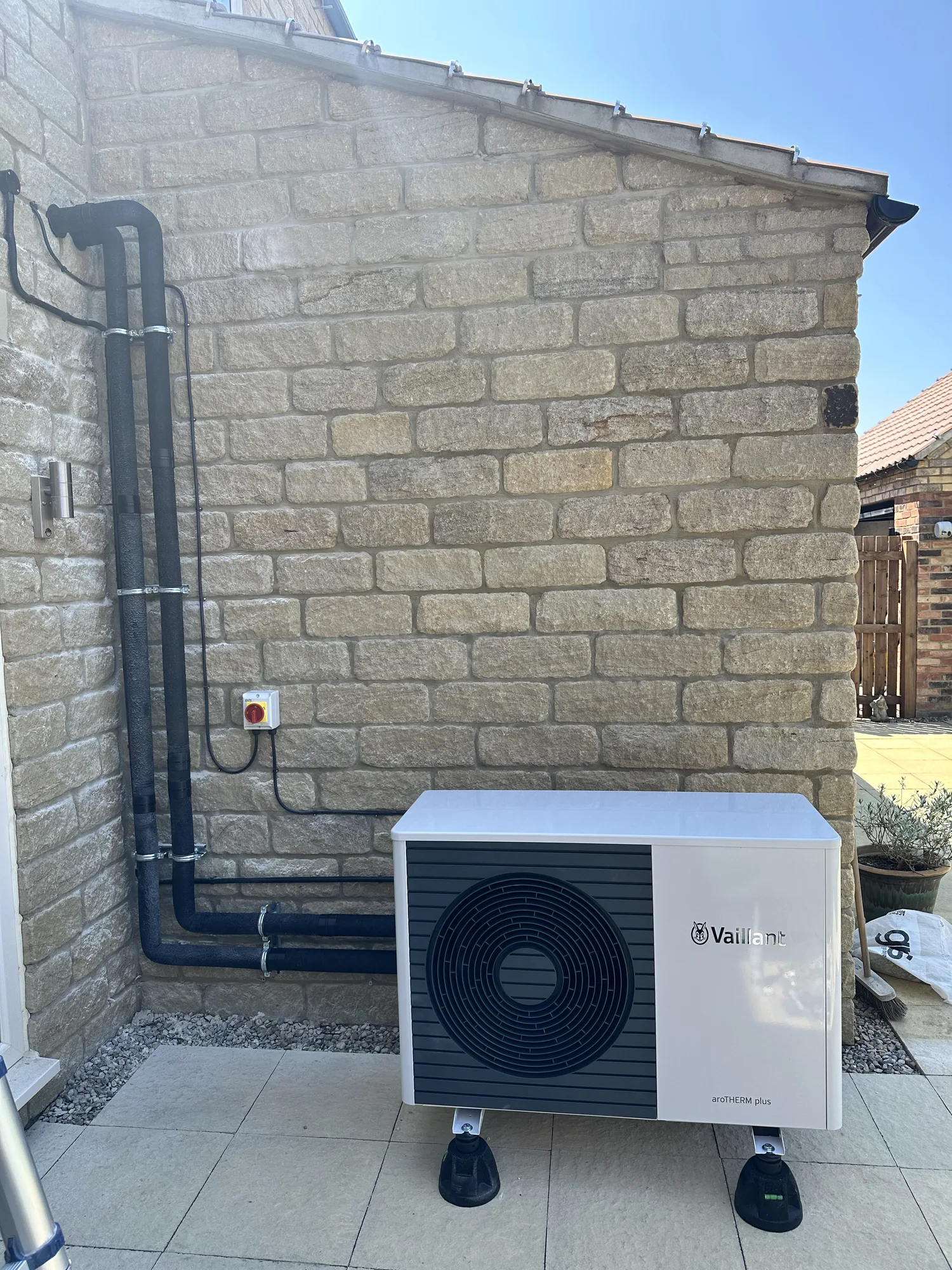 Vaillant air source heat pump outdoor unit against a stone-clad wall with black pipework